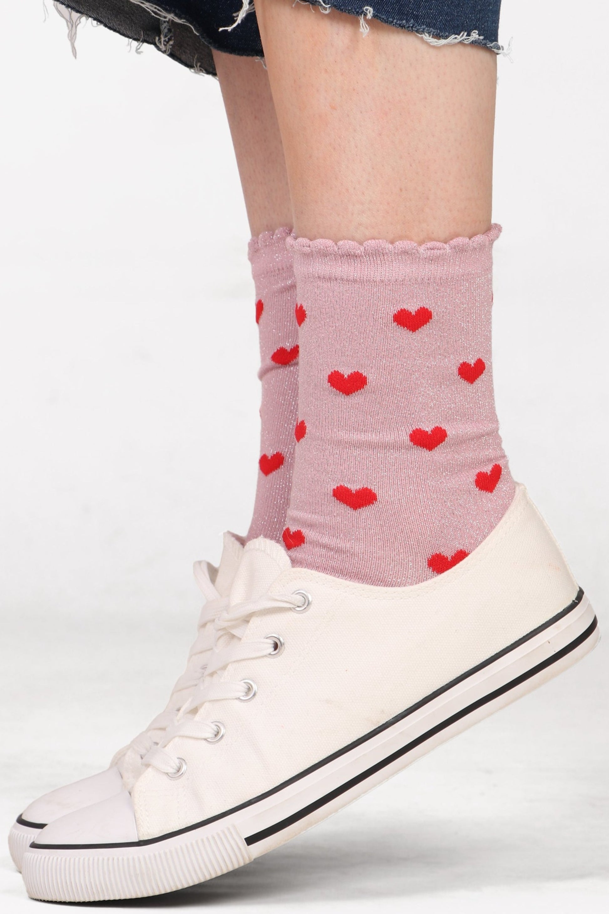 Women's model feet wearing pink glitter heart socks with scallop edge detail. Wearing with white trainers