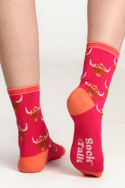 Ladies feet posed facing away from camera showing sole of pink highland cow print socks. Orange contrasting heel can be seen along with sock talk pritned logo on sole of sock