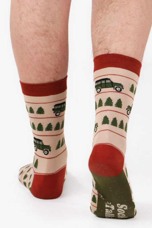 Men's feet walking away from camera wearing sock talk bamboo socks. Socka have off road truck cars printed on them with trees. Socks ar in cream, orange and green tones. Model is walking away from camera to show sock talk logo on sole of sock
